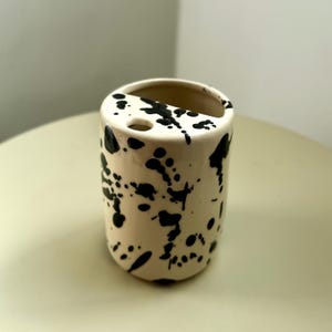 Puede incluir: Un recipiente cilíndrico de cerámica con una base de color blanco roto y un patrón de salpicaduras de pintura negra. La superficie superior presenta una gran abertura semicircular y un orificio circular más pequeño integrados.
