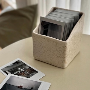 Op de afbeelding: Een gespikkelde beige keramische fotohouder gevuld met instant foto's. Twee instant foto's liggen op de tafel voor de houder. De houder heeft een vierkante basis en schuine zijkanten.