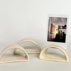 May include: Three ceramic photo holders in soft pastel colors. One is light pink, and the other two are cream-colored. A polaroid photo is displayed in one of the holders. The holders have an arched design.