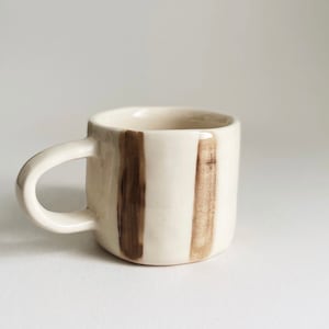 Pode incluir: Uma pequena caneca de cerâmica branca com um design listrado marrom. A caneca tem uma alça arredondada.