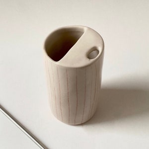 Può includere: Un porta spazzolino da denti in ceramica bianca con strisce rosa. Il porta spazzolino ha due scomparti, uno per uno spazzolino da denti e uno per il dentifricio.