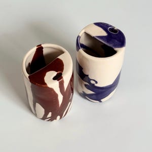 Puede incluir: Dos jarrones de cerámica con base crema y diseños de pintura abstractos. Un jarrón tiene gotas de color marrón oscuro y el otro pintura azul. Cada jarrón tiene una abertura curva y un pequeño agujero.
