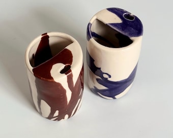 Peut inclure: Deux vases en céramique avec une base crème et des motifs de peinture abstraits. L'un des vases présente des coulures marron foncé, l'autre de la peinture bleue. Chaque vase a une ouverture incurvée et un petit trou.