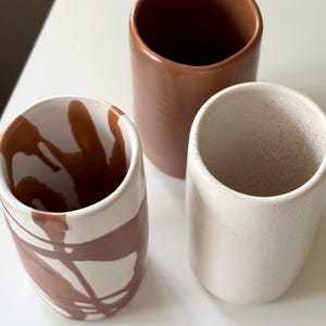 Puede incluir: Tres tazas de cerámica con diferentes diseños. Una taza es marrón con motas blancas, otra es blanca con motas marrones y la tercera es blanca con líneas abstractas marrones.