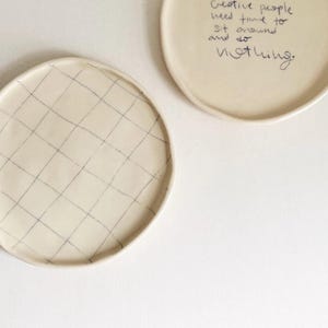 May include: Two off-white ceramic plates. One plate has a grid pattern in dark gray. The other plate has handwritten text that reads, "Creative people need time to sit around and do nothing."