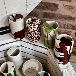 May include: A collection of handmade ceramic vases and small bowls. The vases feature unique designs with colors like white, brown, and green. One bowl has the text "LE MONDE EST UNE POESIE."