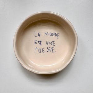 May include: A white ceramic bowl with the French phrase "Le monde est une poésie" written in blue ink.