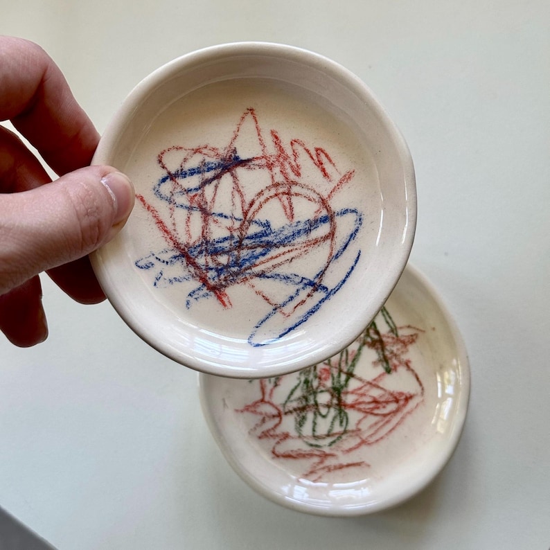 Pu&ograve; includere: Due piatti in ceramica bianca con disegni a cera rossa, blu e verde sulla superficie.