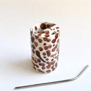 May include: A white ceramic tumbler with brown polka dots, featuring a unique curved opening. A stainless steel straw is placed next to the tumbler. The tumbler is designed for beverages.