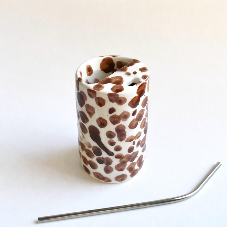 May include: A white ceramic tumbler with brown polka dots, featuring a unique curved opening. A stainless steel straw is placed next to the tumbler. The tumbler is designed for beverages.