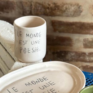 May include: A white ceramic cup and oval plate with black handwritten text. The cup reads "LE MONDE EST UNE POESIE." The plate has similar text. The items are on a white surface, with a brick wall in the background. The cup is cylindrical with a slightly flared top.