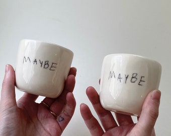 May include: Two white ceramic mugs with the word "MAYBE" written on them in black marker.