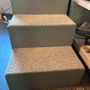 May include: A three-tiered pet step in a light gray color. Each step is covered with a cream-colored, textured carpet. The steps are designed to assist small dogs or cats in reaching higher surfaces.