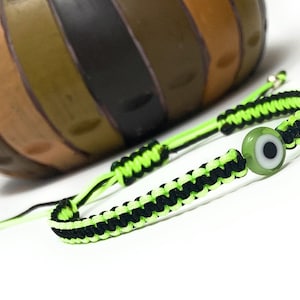 Puede incluir: Una pulsera trenzada verde y negra con un dije de ojo turco verde. La pulsera está atada con un nudo y tiene un cierre de metal plateado.