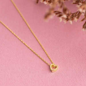 May include: A delicate gold chain necklace with a small, open heart pendant. The necklace is on a pink background.