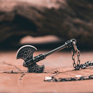 May include: A silver axe pendant with intricate knotwork designs on the blade and handle. The pendant is attached to a silver chain.