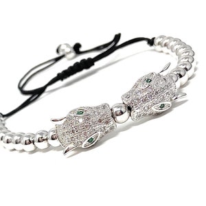 Könnte beinhalten: Ein silbernes Armband mit Perlen und zwei Anhänger in Form von Pantherköpfen. Die Pantherköpfe sind mit klaren Strasssteinen besetzt und haben grüne Augen.