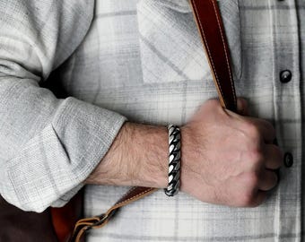 May include: A silver curb chain bracelet is worn on a wrist. The bracelet is made of thick, interlocking links. The person is wearing a gray and white plaid shirt and holding a brown leather strap.