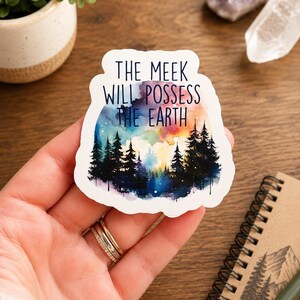 May include: A white sticker with the text "THE MEEK WILL POSSESS THE EARTH" over a watercolor design of a forest and colorful sky. The sticker is held in a hand with gold rings.