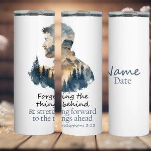 May include: White tumbler with a watercolor design of a man's silhouette with a mountain and trees behind him. The text reads "Forgetting the things behind and stretching forward to the things ahead Philippians 3:13".