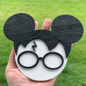 May include: A white wooden ornament shaped like a cartoon character with black ears and hair, wearing round glasses and a lightning bolt on the forehead.