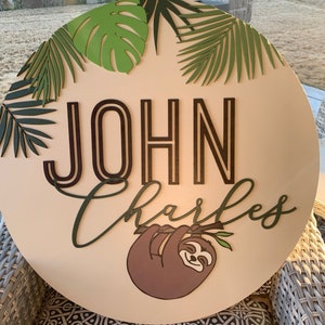 以下が含まれることがあります： 暗い茶色と緑色の文字で「JOHN CHARLES」という名前が書かれた丸い木製の看板。看板にはナマケモノのイラストと緑の葉のアクセントが描かれています。看板は緑と明るい緑の葉に囲まれています。