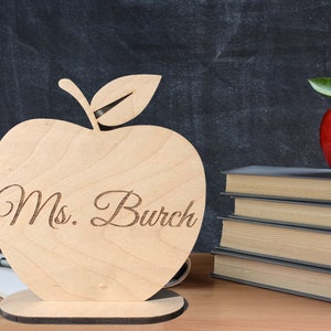 May include: Wooden apple-shaped teacher appreciation gift with the text "Ms. Burch" engraved on the front. The apple is sitting on a wooden base and is in front of a stack of books.