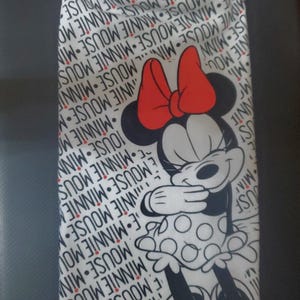 May include: White hand towel featuring a Minnie Mouse graphic. The towel has a black fringe at the top and a repeating pattern of "MINNIE MOUSE" in black. Minnie Mouse is depicted in black, white, and red.