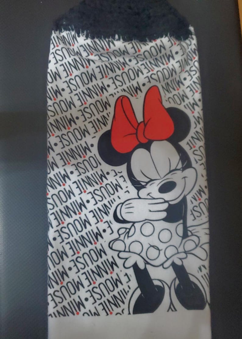 May include: White hand towel featuring a Minnie Mouse graphic. The towel has a black fringe at the top and a repeating pattern of "MINNIE MOUSE" in black. Minnie Mouse is depicted in black, white, and red.