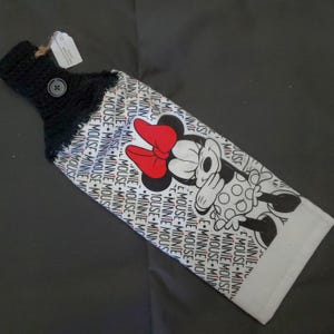 May include: A black and white Minnie Mouse kitchen towel with a crocheted top and button closure. The towel features a repeating pattern of "MINNIE MOUSE" and a large image of Minnie Mouse with a red bow.