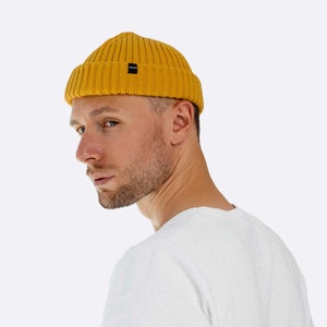 Könnte beinhalten: Eine Person trägt eine senfgelbe Mütze mit einem schwarzen Logo an der Seite. Die Mütze ist aus geripptem Strickstoff gefertigt. Die Person trägt ein weißes T-Shirt.