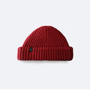 Puede incluir: Un gorro de punto rojo con una pequeña etiqueta negra que dice "ORIGINAL".