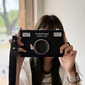 May include: Black camera-shaped bag with a long black strap. The bag has a large lens design and the text "FILWHITE BAG X-BAIDA 1030". The bag has a silver-colored accent on the top left corner.
