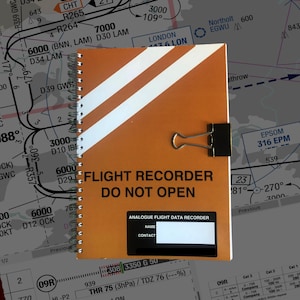 May include: An orange spiral-bound notebook with the text "FLIGHT RECORDER DO NOT OPEN" and white diagonal stripes. A black clip secures the notebook. The notebook includes a label for "NAME" and "CONTACT".