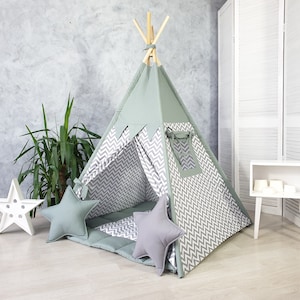Puede incluir: Una tienda de campaña tipi para niños con estampado de chevron verde y gris, una alfombra de suelo con estampado de chevron gris y blanco y dos almohadas grises.