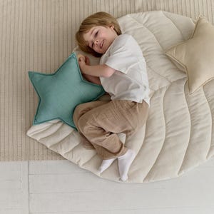 May include: A child lays on a cream-colored quilted leaf-shaped play mat with two star-shaped pillows. One pillow is teal and the other is beige.
