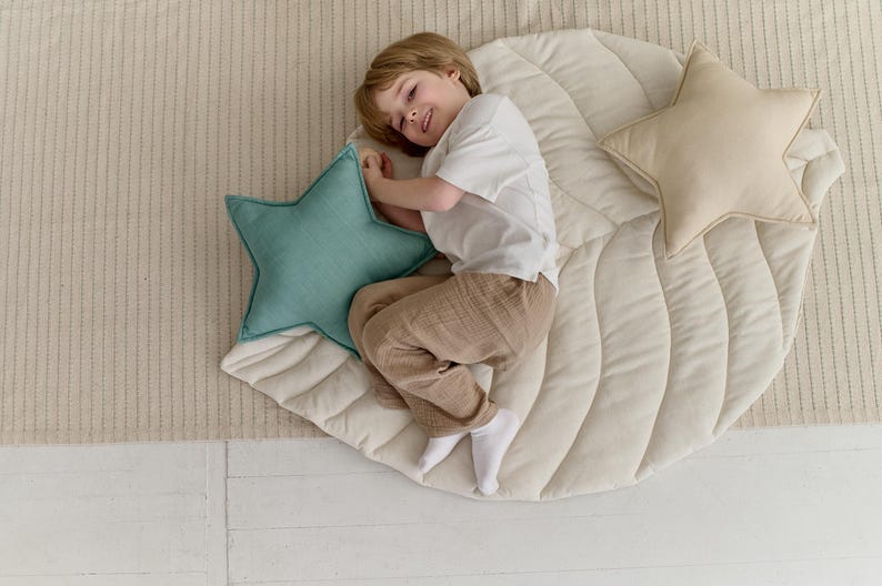May include: A child lays on a cream-colored quilted leaf-shaped play mat with two star-shaped pillows. One pillow is teal and the other is beige.