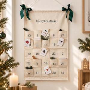 Può includere: Un calendario dell'avvento da parete in tessuto color crema con la scritta Merry Christmas stampata in alto. Presenta venticinque tasche individuali, ciascuna contrassegnata da un numero nero, ed è appeso a un cordino con nastri verde scuro.