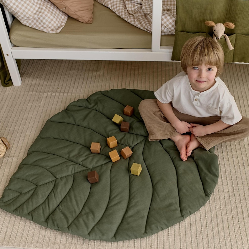 May include: A green leaf-shaped play mat with a quilted texture. A child is sitting on the mat playing with wooden blocks.