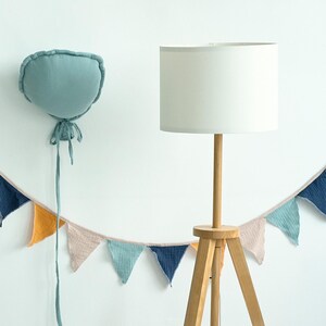 May include: A white lampshade with a wooden tripod stand and a light blue fabric balloon with a string hanging from it. A garland of colorful fabric triangles hangs below the lamp.