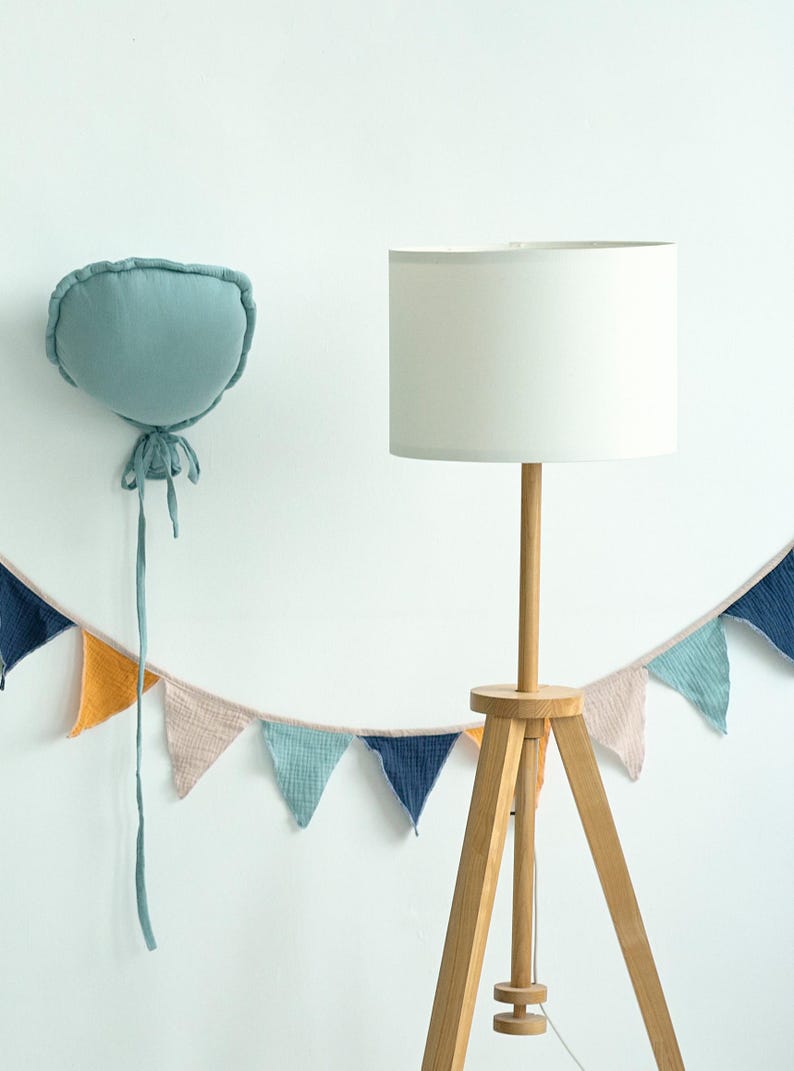 May include: A white lampshade with a wooden tripod stand and a light blue fabric balloon with a string hanging from it. A garland of colorful fabric triangles hangs below the lamp.