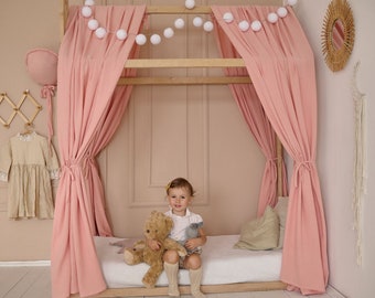 May include: A young girl sits on a bed with pink curtains and a white bedspread. She is holding two stuffed animals, a brown bear and a gray bunny. The bed is decorated with a string of white pom-poms.