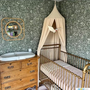 May include: A gold metal cot with a white canopy and a white bird mobile hanging above it. The cot is in a room with green floral wallpaper and a wooden chest of drawers with a white changing mat on top.