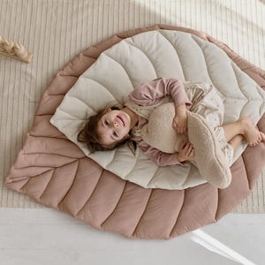 May include: A child lays on a large, pink and cream leaf-shaped play mat. The mat is quilted and has a soft, plush texture. The child is holding a plush toy and smiling.