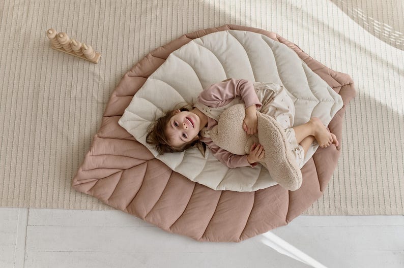 May include: A child lays on a large, pink and cream leaf-shaped play mat. The mat is quilted and has a soft, plush texture. The child is holding a plush toy and smiling.