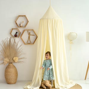 May include: A white cotton canopy tent for a child's bedroom. The tent is draped over a wooden frame and has a white fabric lining. A young girl is standing inside the tent, looking out at the camera. The tent is decorated with a stuffed animal and a heart-shaped balloon.