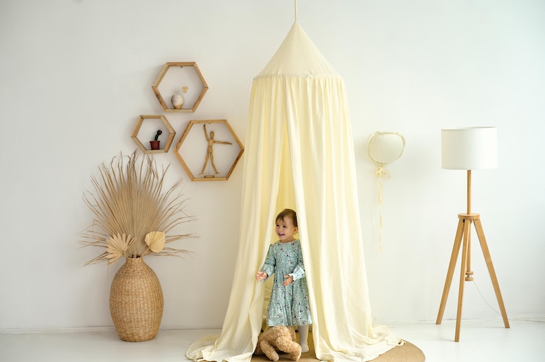 May include: A white cotton canopy tent for a child's bedroom. The tent is draped over a wooden frame and has a white fabric lining. A young girl is standing inside the tent, looking out at the camera. The tent is decorated with a stuffed animal and a heart-shaped balloon.