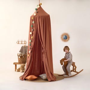 May include: A terracotta-colored fabric canopy with a string of colorful pom-poms hanging from the top. A child is sitting on a wooden rocking horse in front of the canopy.