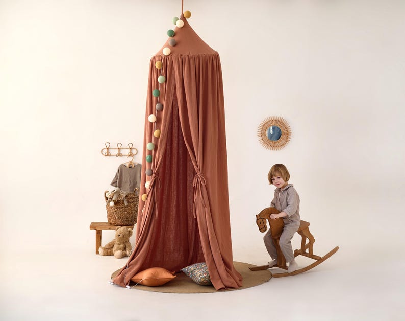May include: A terracotta-colored fabric canopy with a string of colorful pom-poms hanging from the top. A child is sitting on a wooden rocking horse in front of the canopy.