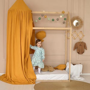 May include: A young girl in a blue and white floral dress stands inside a wooden playhouse with a yellow canopy. The playhouse has a white mattress and a stuffed animal on it. The playhouse is decorated with a string of lights and a wooden mirror.